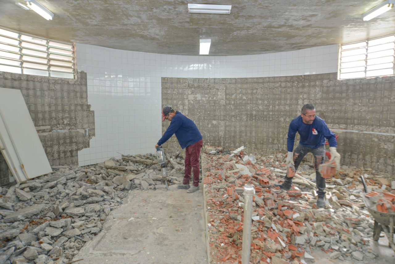 DESCRIÇÃO DA IMAGEM
Dois homens estão trabalhando em uma obra, dentro de um espaço fechado, com janelas basculantes. Parte da parede está com azulejos e a maior parte já está sem. No chão estão resíduos da obra. É uma obra em banheiro.