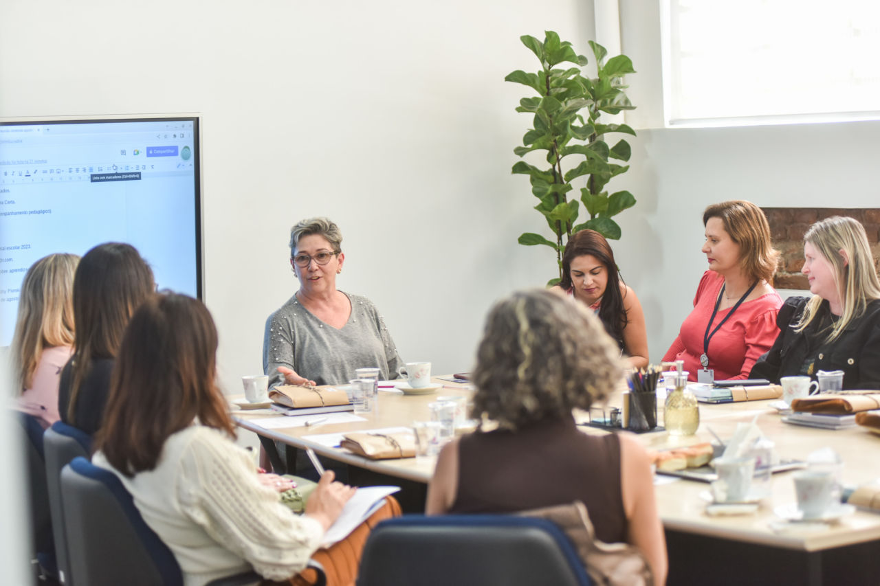 Em uma mesa, estão sentadas 7 mulheres, que aparecem na foto. No foco, está a gestora de Educação. Atrás está a Tela Interativa com a apresentação.