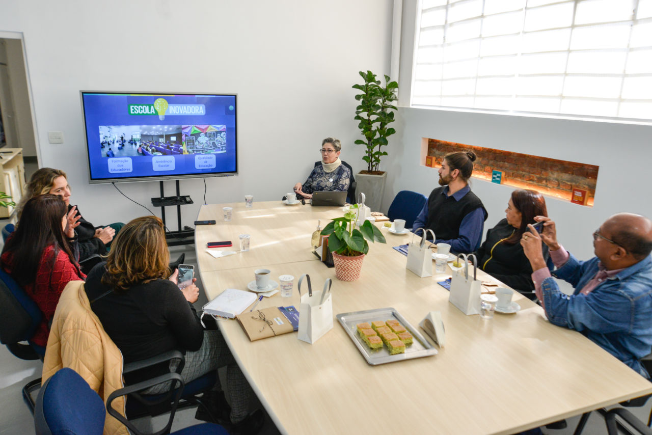 Em uma sala com uma mesa no centro, 7 pessoas estão sentadas em volta da mesa. Em frente, tem uma tela interativa com apresentação da Educação de Jundiaí, o logo Escola Inovadora está no foco.