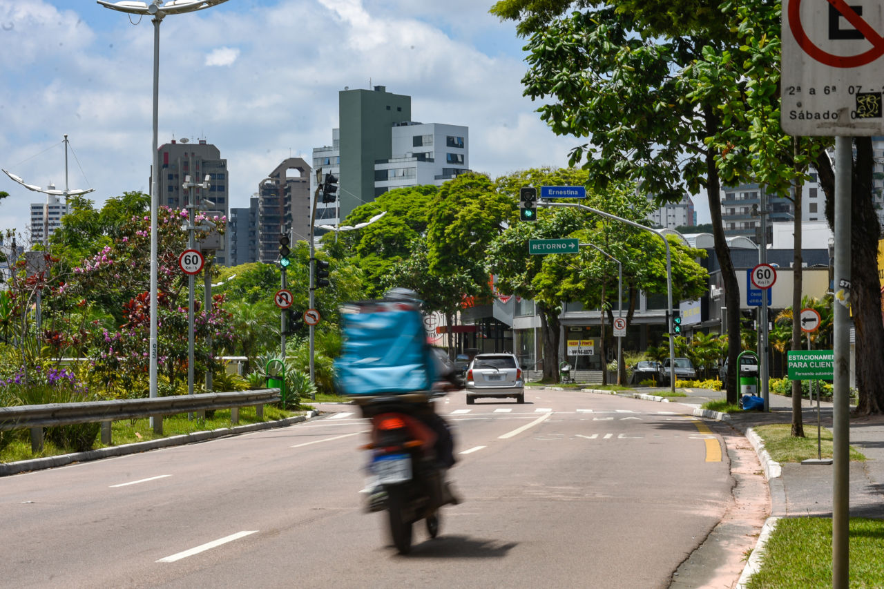DESCRIÇÃO DE IMAGEM: foto feita em curva de avenida, com imagem de motociclista em primeiro plano, na faixa da direita e, ao fundo, carro, na faixa da esquerda