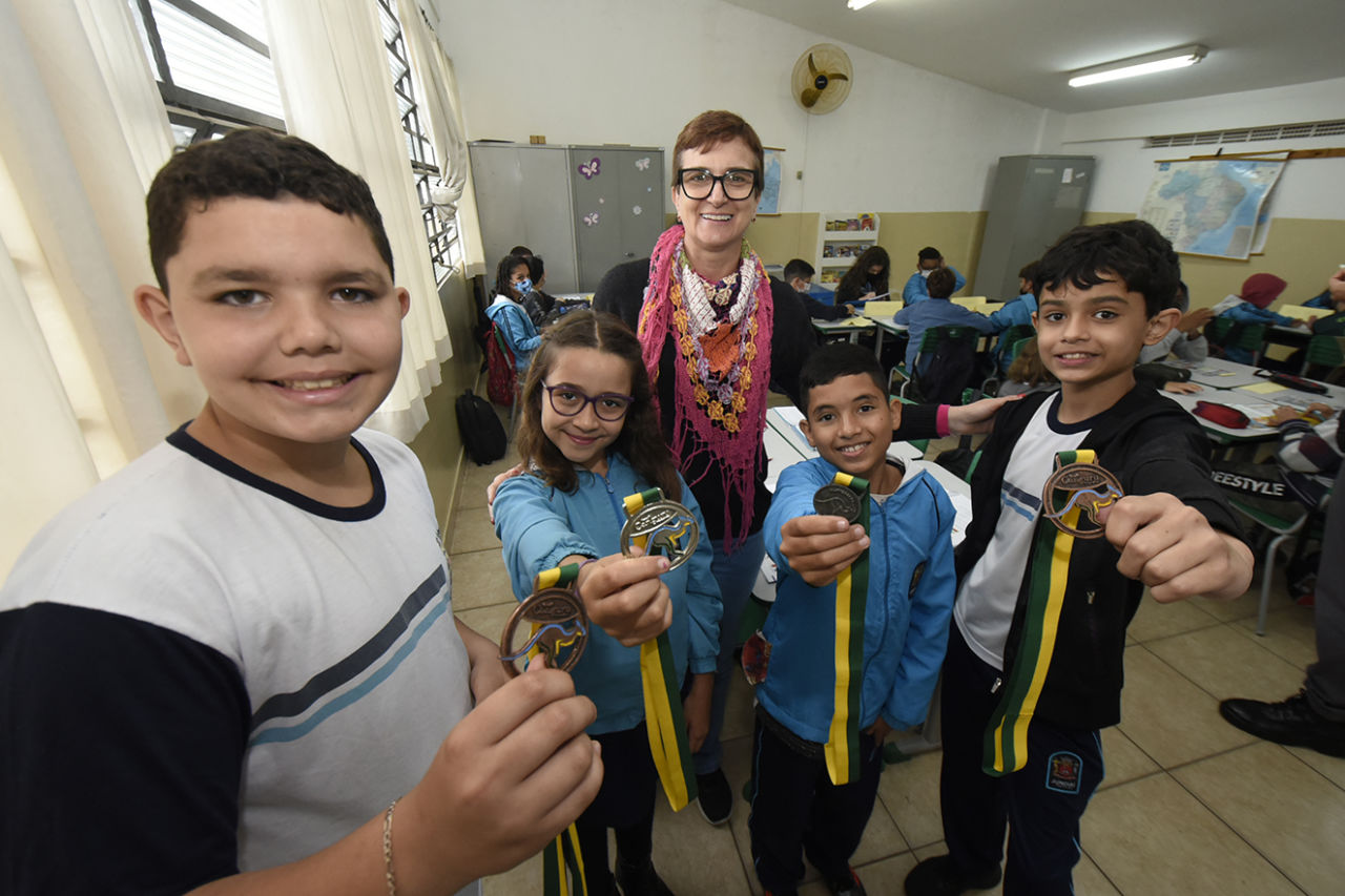 Nesta imagem contém os quatro alunos medalhistas com a professora de matemática, eles estão na sala de aula, de pé e mostrando a medalha para a câmera. 