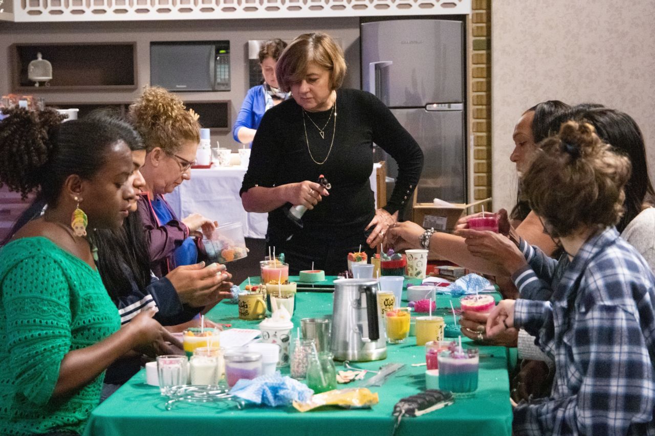 DESCRIÇÃO DE IMAGEM: pessoas sentadas ao redor de mesa de trabalho, confeccionando velas artesanais em embalagens diversas, como copos, xícaras entre outros