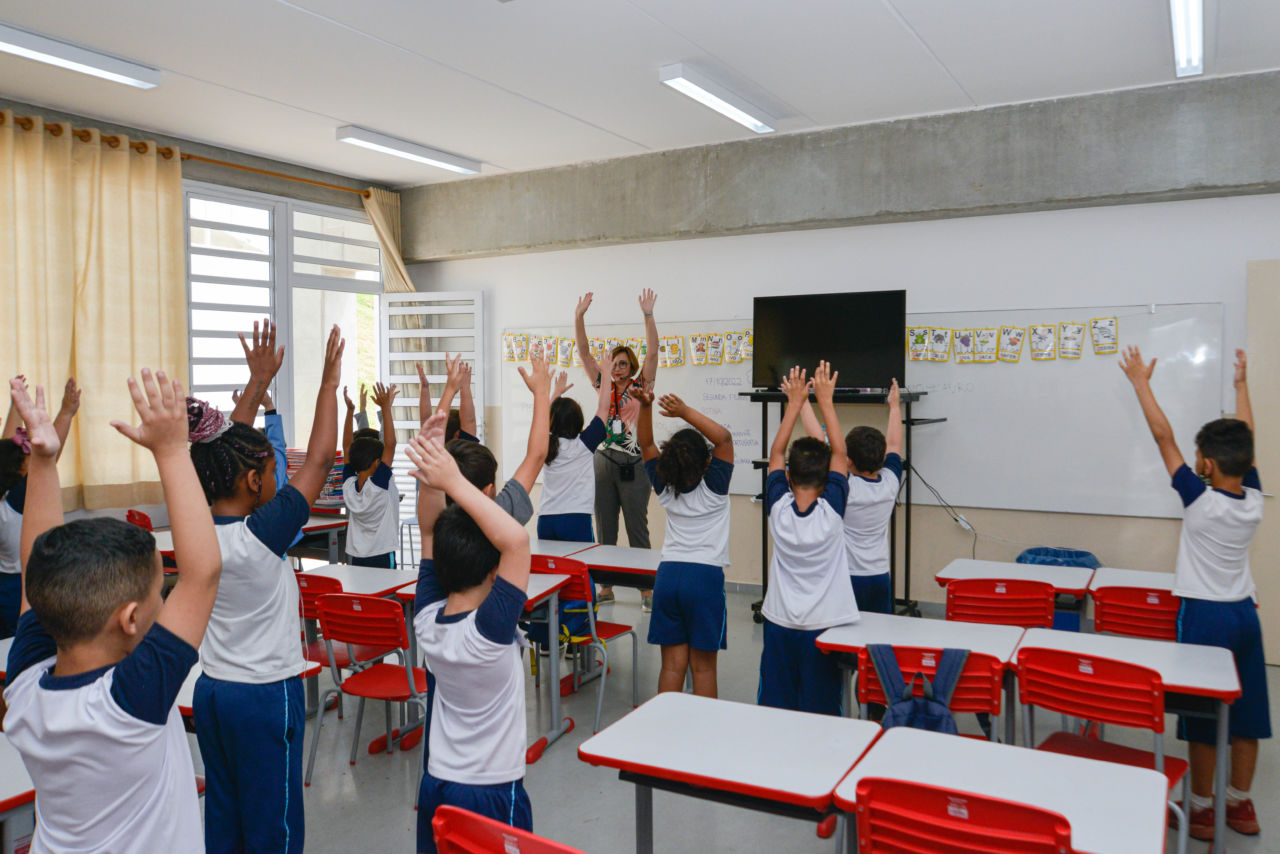 Nesta imagem contém alunos em sala de aula com os braços erguidos, eles estavam fazendo a coreografia da música.