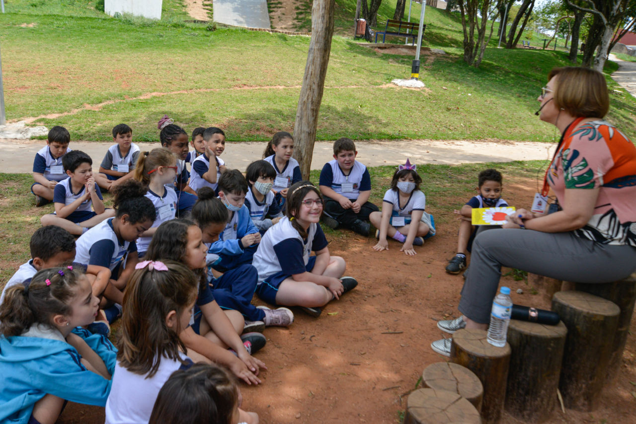 Nesta imagem contém alunos sentados no chão do bosque da escola e a professora a frente deles explicando a atividade
