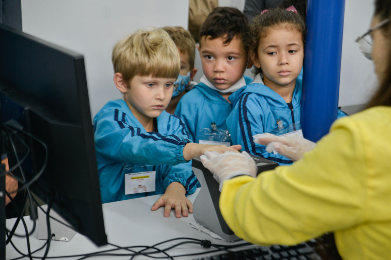 Na imagem contem três crianças observando seu colega no momento da coleta de digitais, um dos passos para a retirada do RG