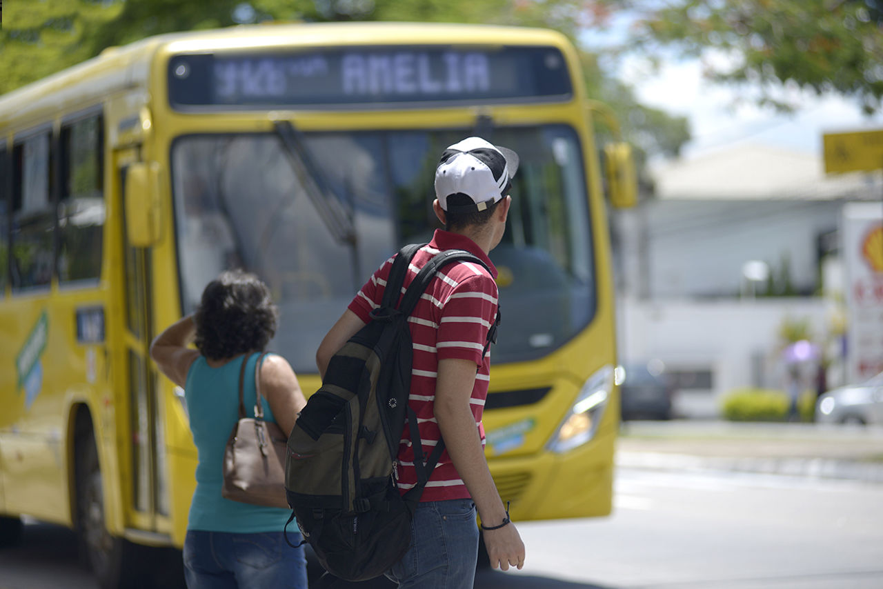 DESCRIÇÃO DE IMAGEM: rapaz e mulher aguardam ônibus, de costas, olhando para a rua, com ônibus de transporte público de Jundiaí ao fundo