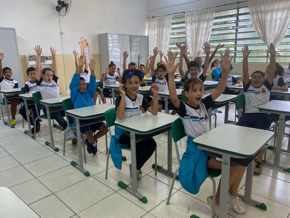 Nesta imagem contem alunos em sala de aula com os braços levantados, comemorando uma conquista. 