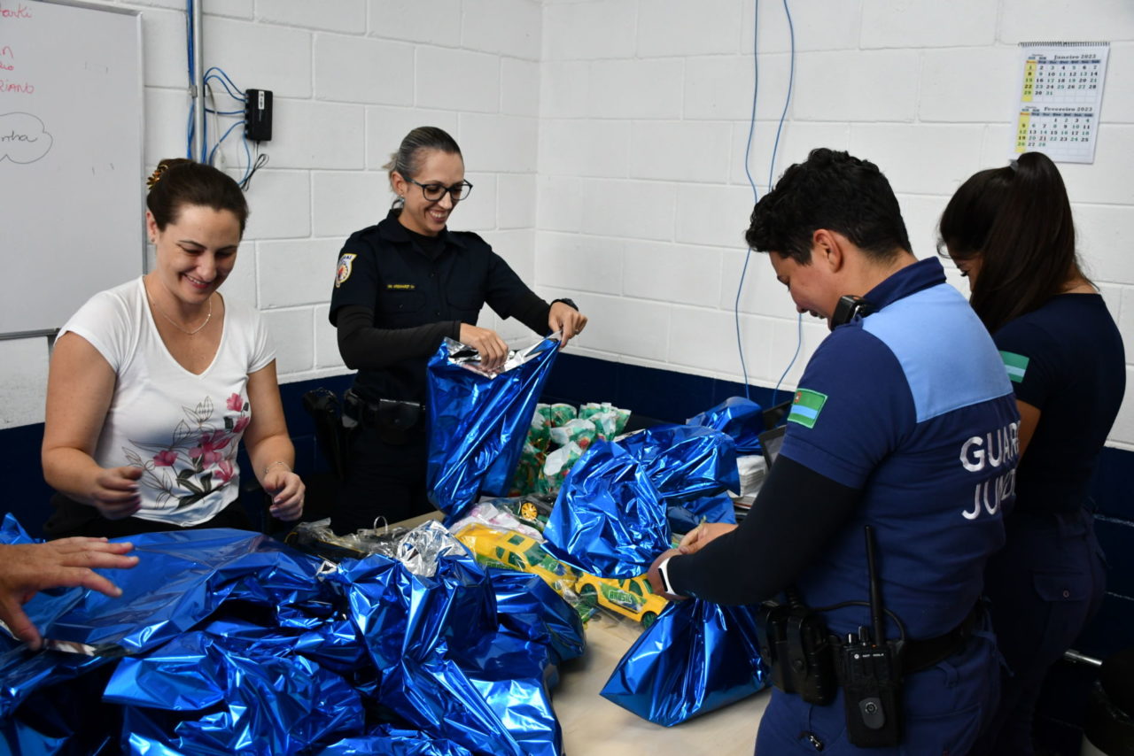 DESCRIÇÃO DA IMAGEM: no entorno de uma mesa com brinquedos e pacotes de embrulho de presente azul metálico, quatro guardas municipais mulheres empacotam os brinquedos com sorrisos nos rostos