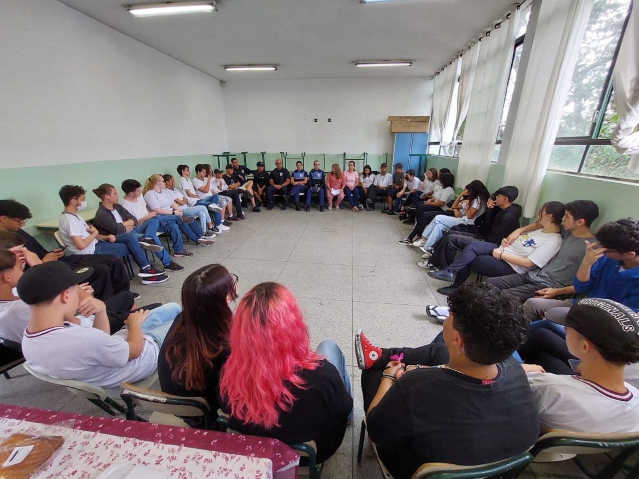 DESCRIÇÃO DE TEXTO:  alunos sentados em cadeiras formando um círculo recebem orientações de Guardas Municipais sobre violência doméstica