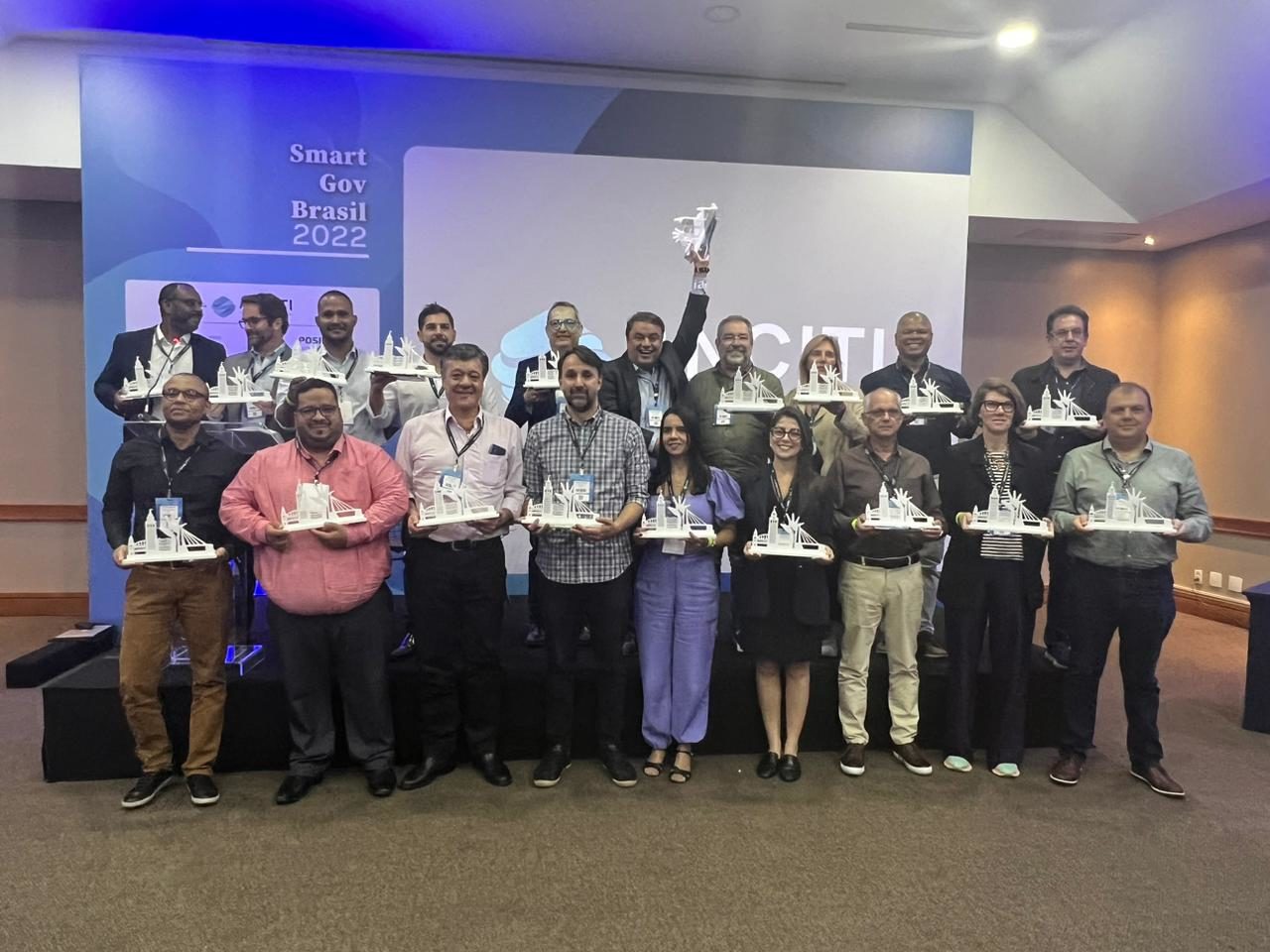 DESCRIÇÃO IMAGEM: foto com várias pessoas representantes das cidades premiadas, seguram troféu. Enfileirados em frente a painel do evento Smart Gov Brasil 2022