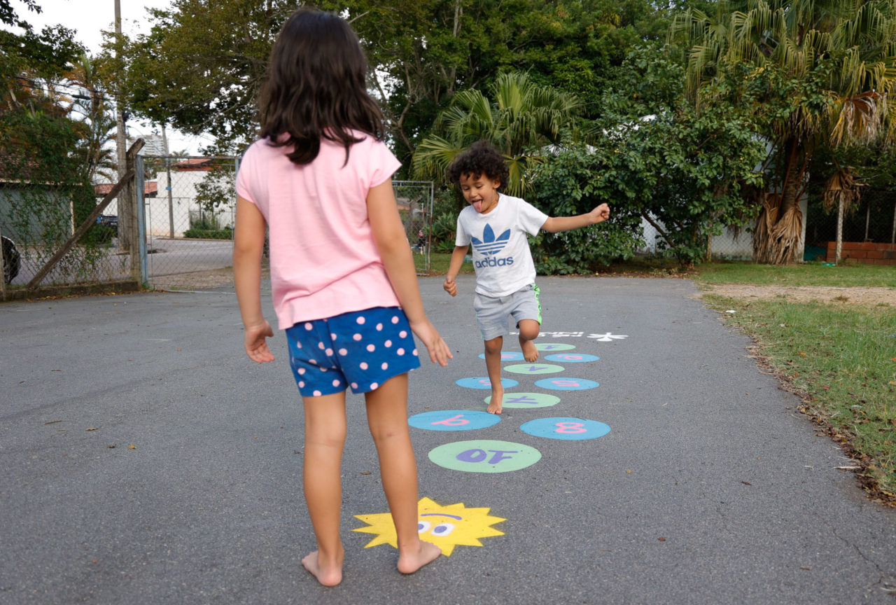 DESCRIÇÃO DA IMAGEM
Duas crianças brincam de amarelinha em brincadeira pintada no solo