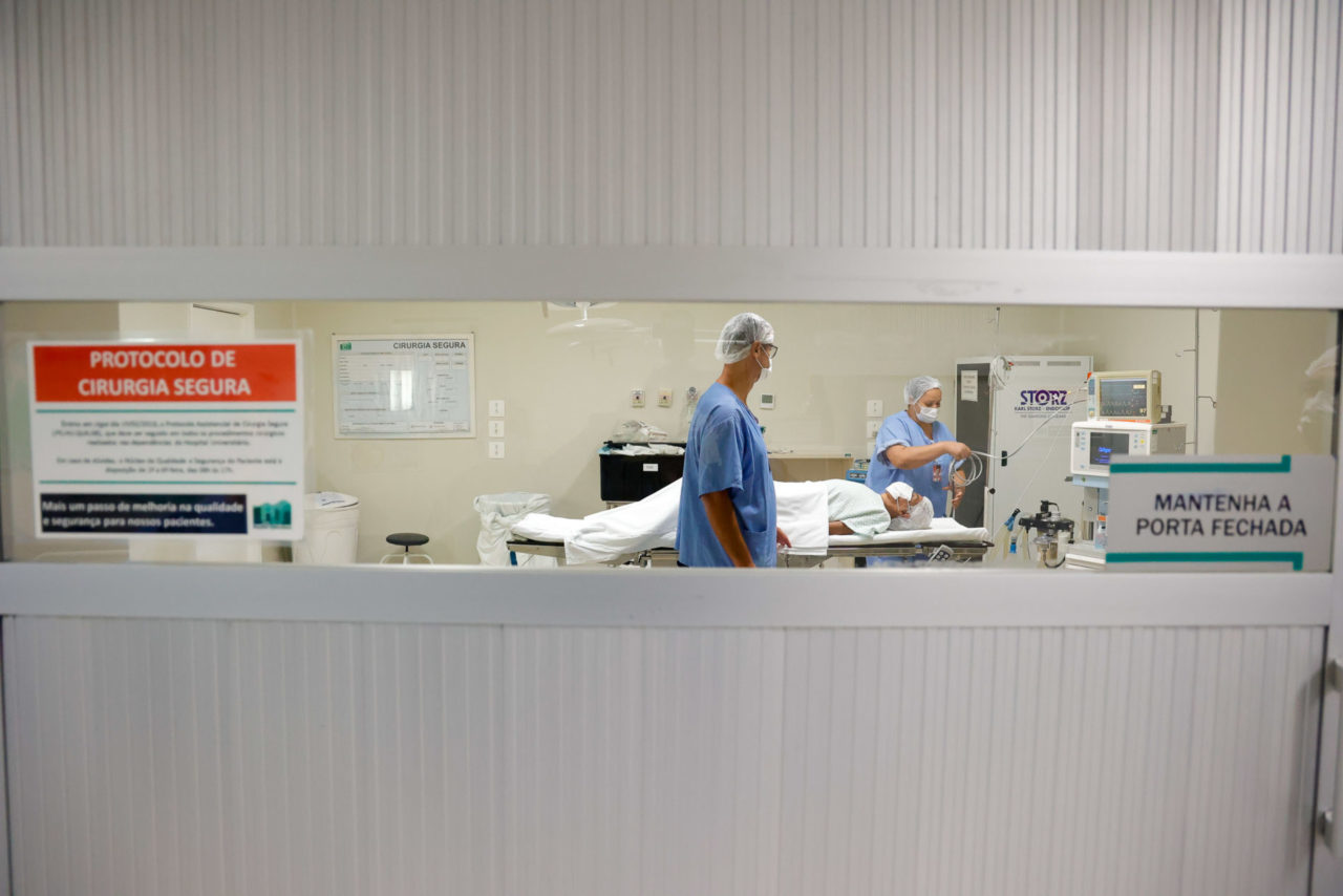 DESCRIÇÃO DA IMAGEM: imagem de porta de sala de cirurgia, com visor de vidro, que apresenta a sala de procedimentos, com paciente deitado em maca e profissionais paramentados no entorno fazendo o preparo para a cirurgia