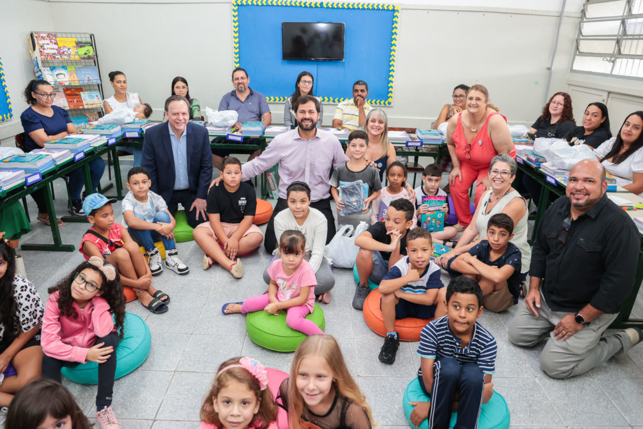 DESCRIÇÃO DA IMAGEM
Em uma sala de aula há adultos e nas carteiras e crianças e adultos sentados no chão.