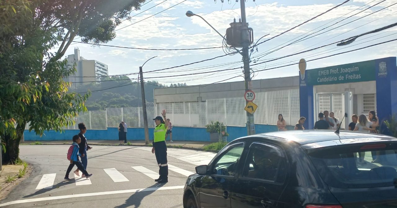 Na foto, a agente de trânsito está com o braço esquerdo esticado para sinalizar para o motorista parar enquanto mãe e filha atravessam a rua em frente à escola.