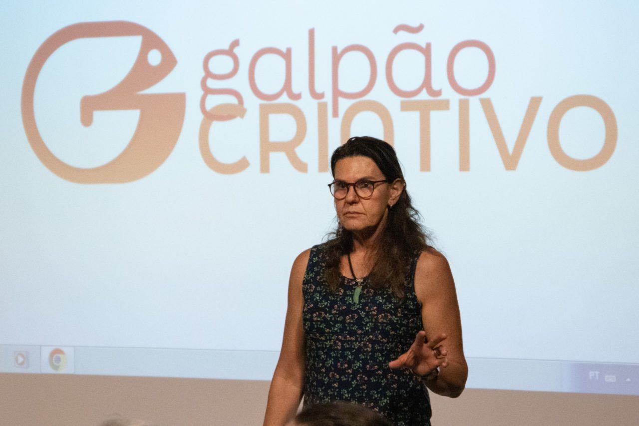 DESCRIÇÃO DE IMAGEM: foto de supervisora Monica Gropelo, à frente de painel com projeção de logomarca Galpão Criativo. 