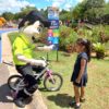 Na foto, o mascote da Unidade de Gestão de Mobilidade e Transporte, Vagninho, brinca com uma menina que está de bicicleta, e sorrindo.