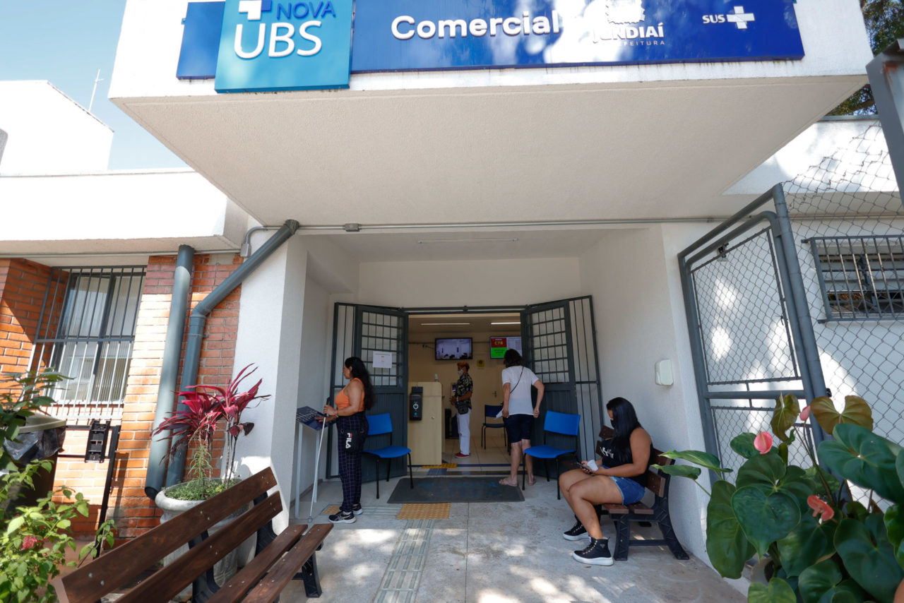 DESCRIÇÃO DE IMAGEM: entrada da 
Nova UBS Comercial, com placa onde está escrito o nome da unidade, abaixo, bancos para as pessoas sentarem e aguardarem a entrada na unidade