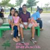 Na imagem, estão os pais da pequena Mariana, que está prestes a completar 3 anos, e estão sorrindo enquanto aguardam o o ônibus, sentados em um banco de concreto verde e com colorido da Cidade das Crianças.