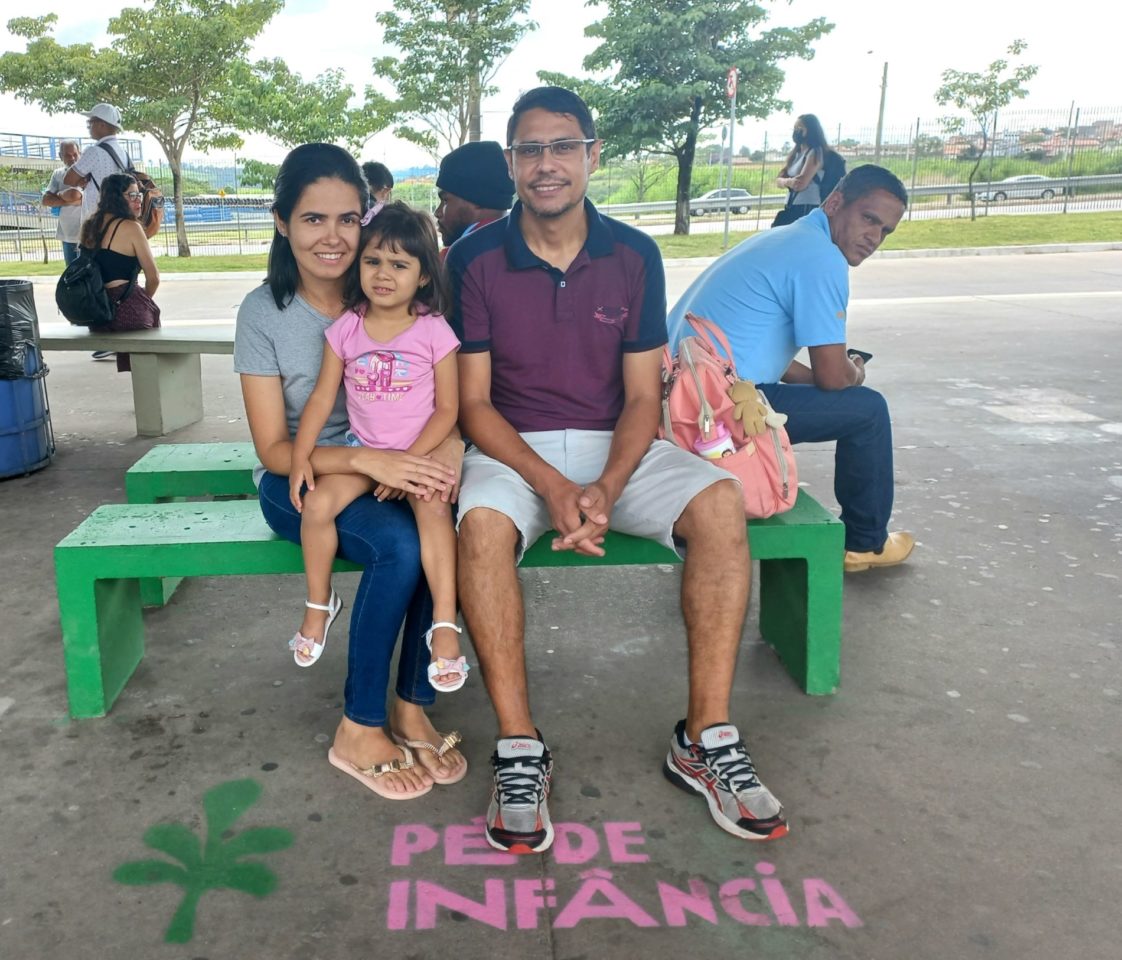 Na imagem, estão os pais da pequena Mariana, que está prestes a completar 3 anos, e estão sorrindo enquanto aguardam o o ônibus, sentados em um banco de concreto verde e com colorido da Cidade das Crianças.