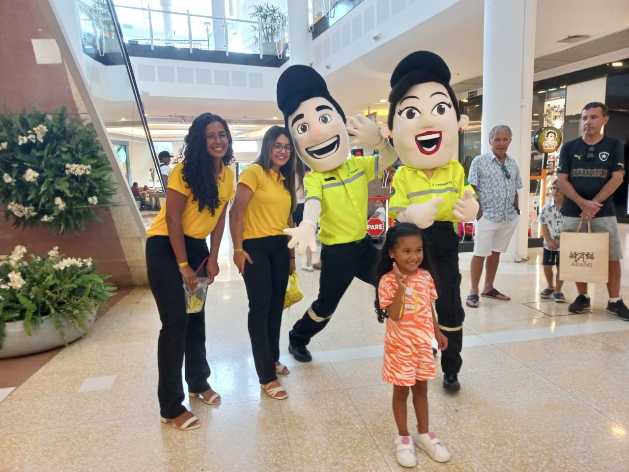 Na imagem, os bonecos mascotes de Mobilidade e Transporte, chamados Vaguinho e Julinha, estão no corredor de um shopping em Jundiaí, com uma criança, a Myrela, que está sorrindo.
