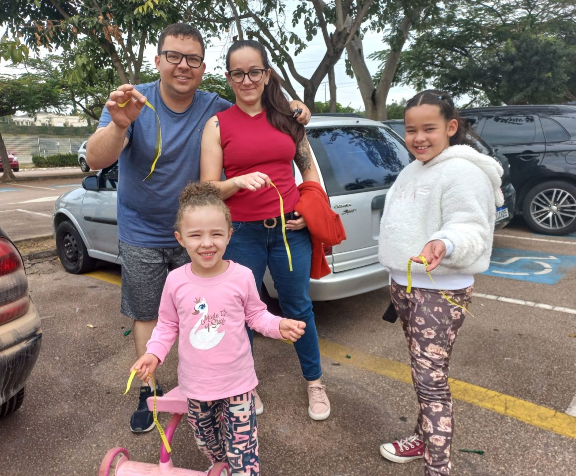 Na imagem, estão o Christian, com a esposa e duas filhas no Parque da Cidade. Eles seguram e mostram as fitas que ganharam na ação educativa do Maio Amarelo.