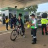 Na imagem, mulher ciclista com capacete recebe uma fita amarela escrito Trânsito Seguro da equipe da Unidade de Gestão de Mobilidade e Transporte, na portaria do Parque da Cidade.