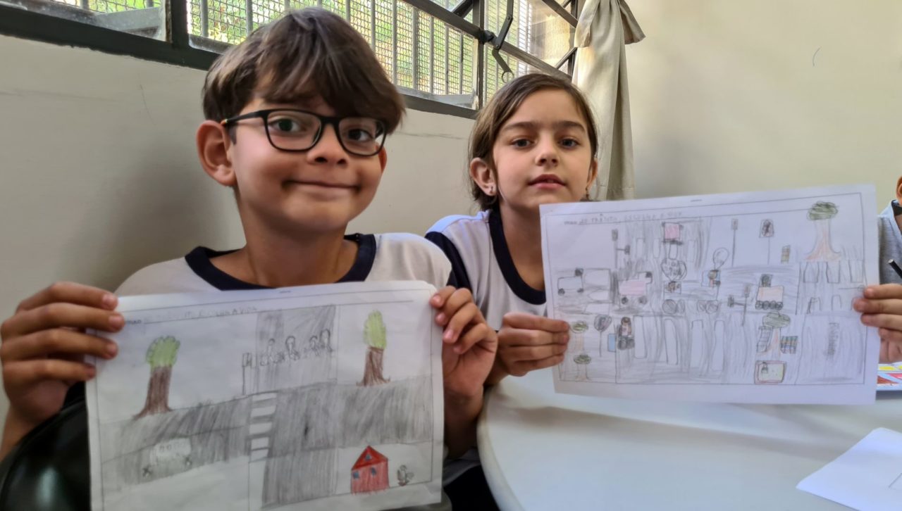 Na imagem, os alunos Henry e Giovanna mostram os desenhos, com uma rua, casas e comércios, árvores e pessoas. Eles estão no segundo ano da EMEB Prof.ª Marina de Almeida Rinaldi Carvalho, no Jardim das Tulipas.