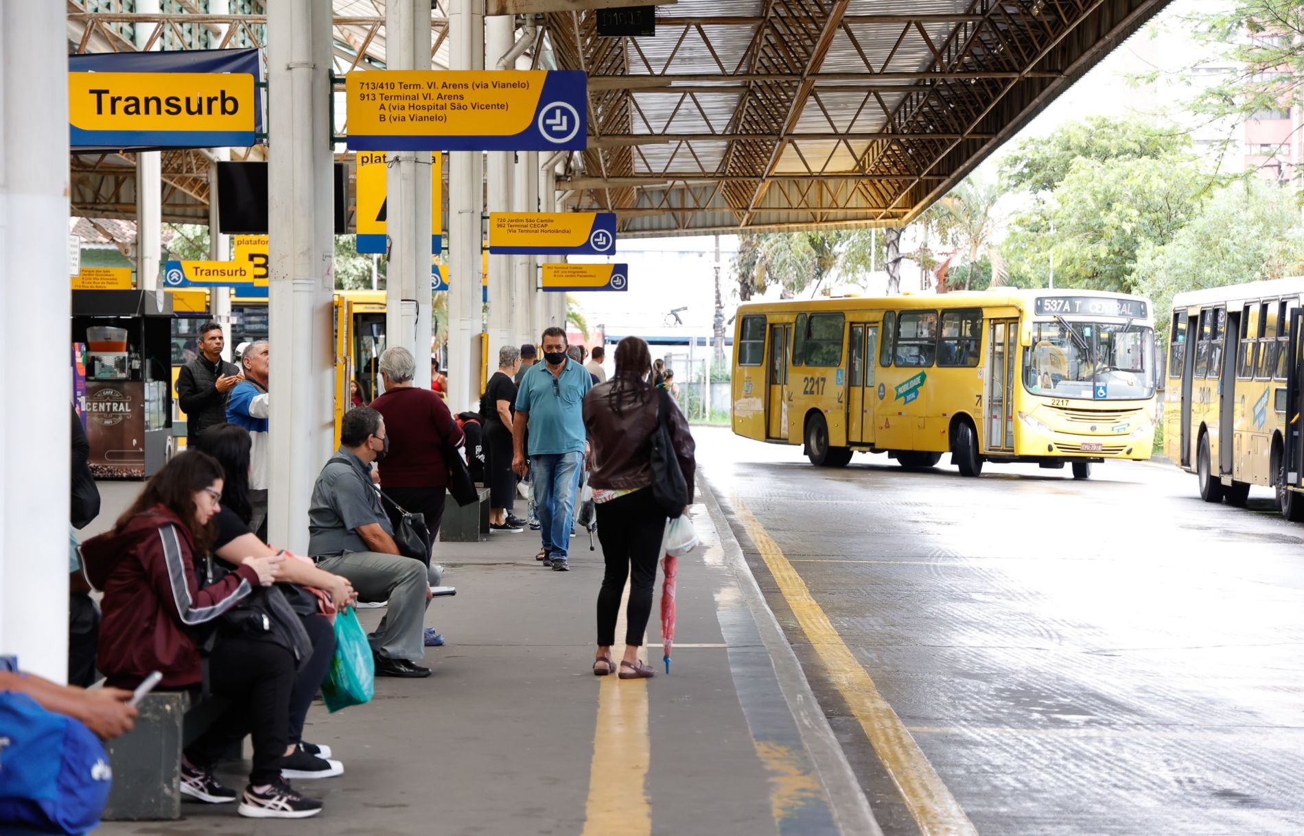 Na imagem, está o terminal de ônibus central com pessoas aguardando na plataforma, e alguns ônibus saindo para fazer os percursos.