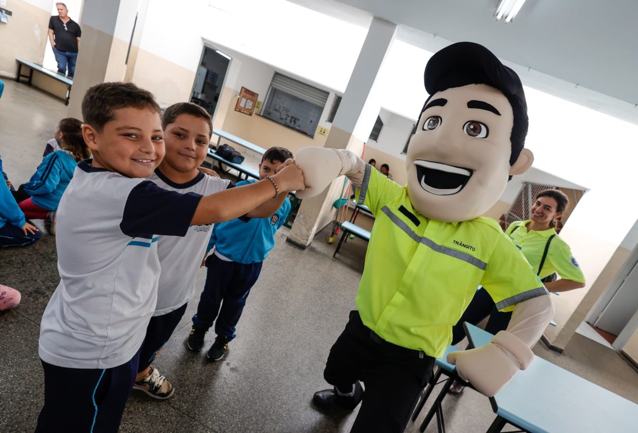 Na imagem, o mascote Vaguinho dá as mãos para os alunos que participaram da ação na EMEB Antonino Messina sobre as regras e equipamentos de segurança de trânsito.