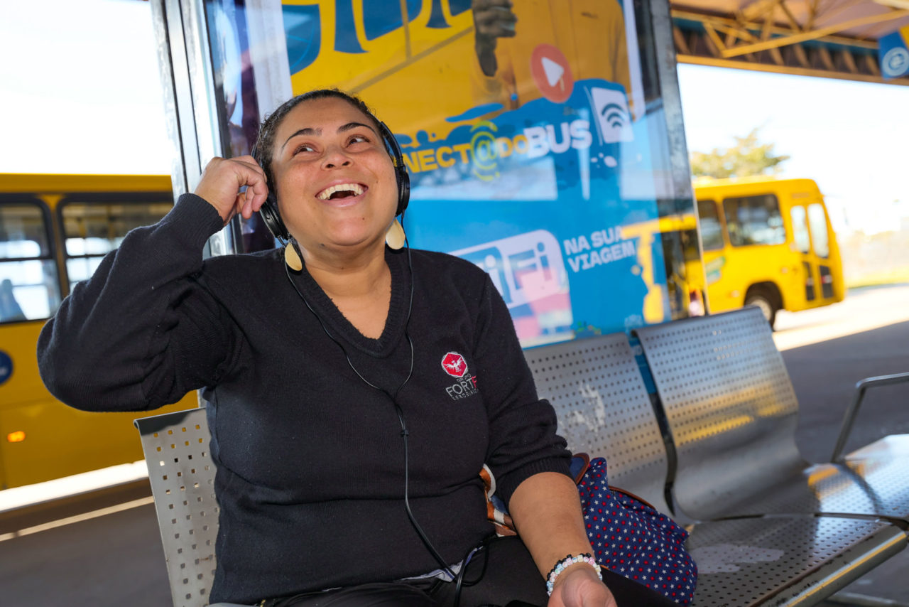Na imagem, tem uma moça, Kamilla, com o fone de ouvido e o celular conectado na internet, e ela contou sorrindo que é ótimo saber que tem wi-fi nos ônibus, porque, ela utiliza ônibus diariamente.