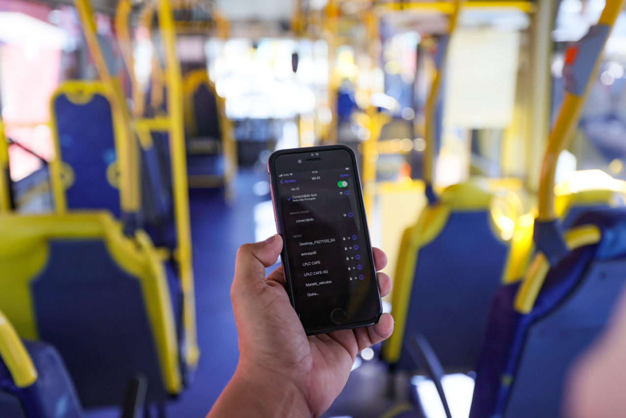Na imagem, tem uma mão segurando um celular, com a tela inicial com o nome da rede de wi-fi nos ônibus: conect@adobus. Os ônibus municipais de Jundiaí contam com sinal de internet de graça para os usuários.
