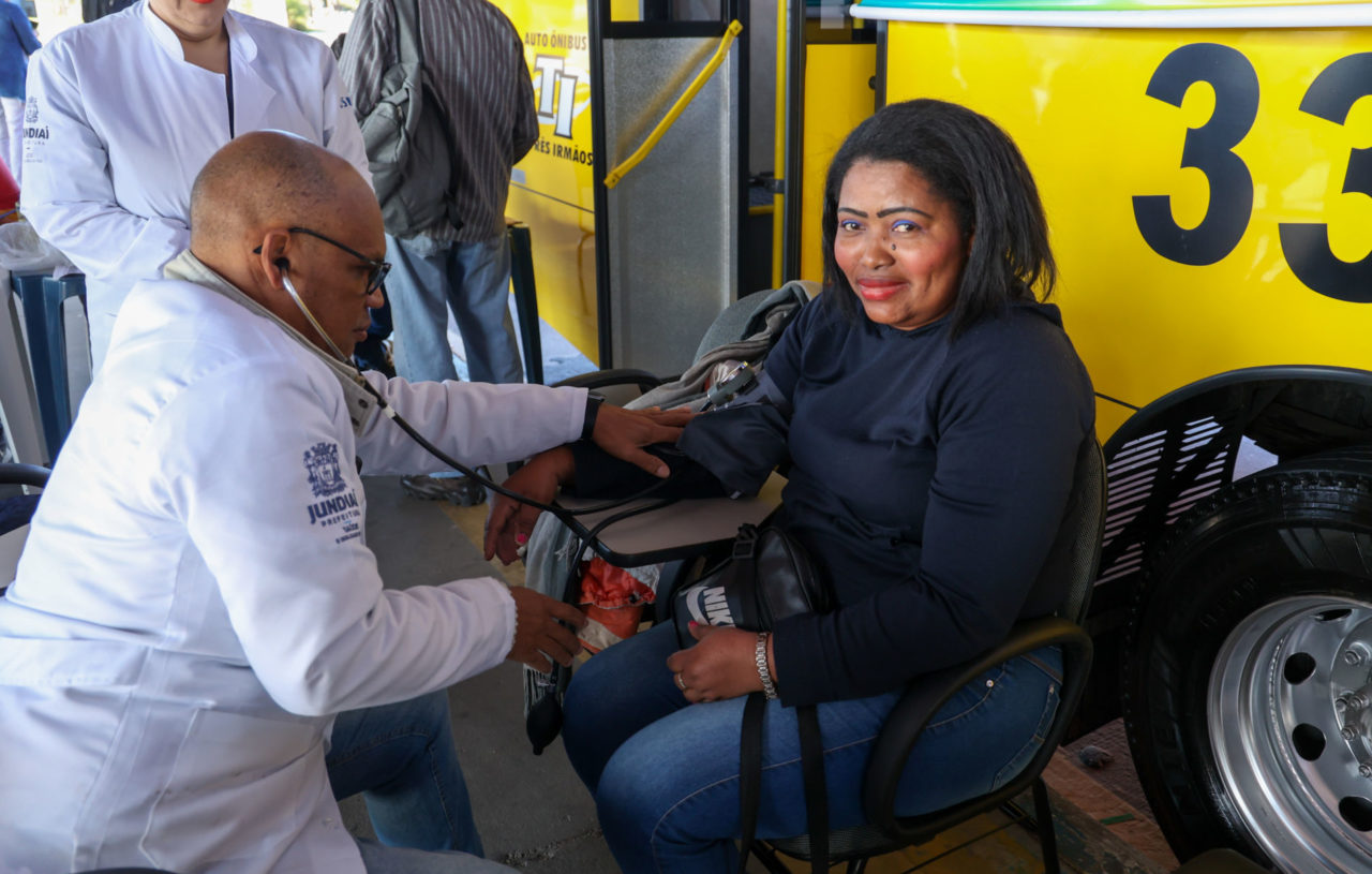 Na imagem, está uma mulher que está passando por atendimento para aferição de pressão arterial. Ela está sentada do lado de fora do ônibus, onde tem uma equipe de enfermeiros.