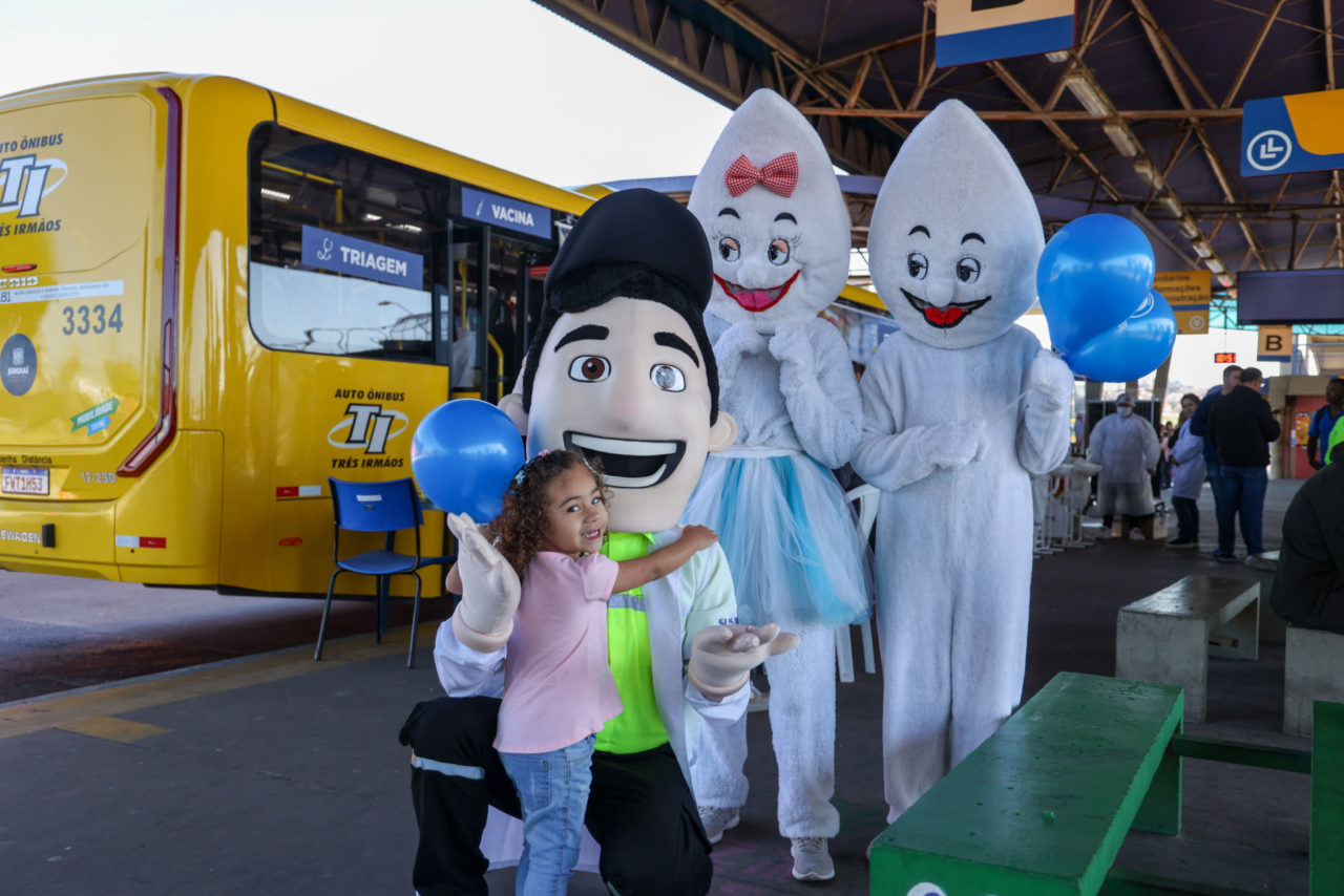 Na imagem, tem uma criança - menina - com os mascotes da Saúde - Zé Gotinha e Maria Gotinha - e o Vaguinho da UGMT