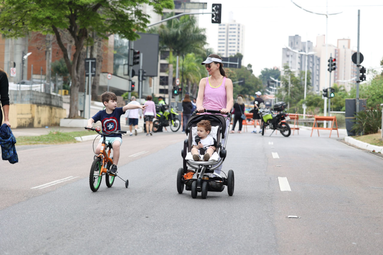 Na foto está uma mulher acompanhada pelo filho em uma bike e outro menor sendo empurrado no carrinho.