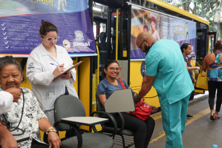 Na imagem estão duas mulheres sentadas, e dois enfermeiros colocam o aparelho de aferição de pressão arterial, enquanto uma outra enfermeira anota os dados e resultados.