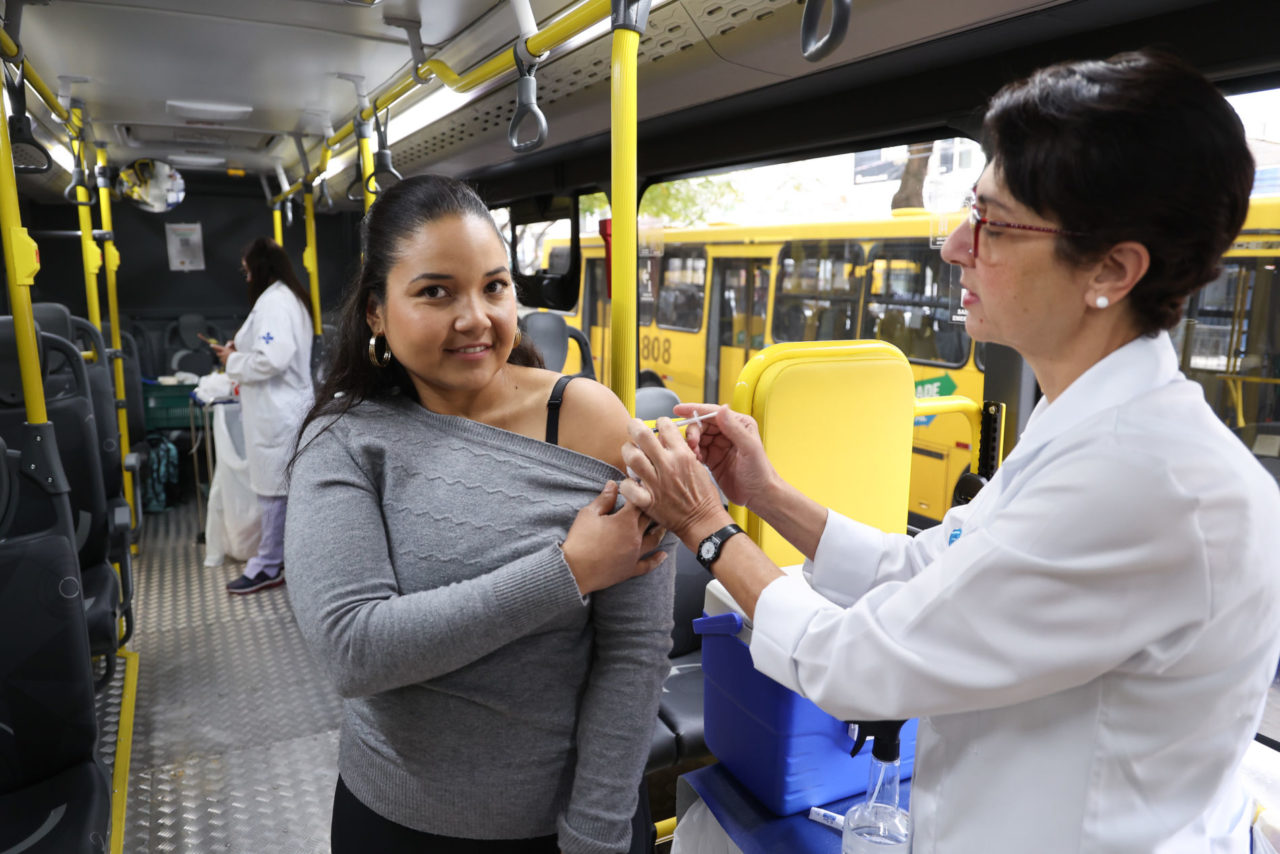 Na imagem está uma mulher com uma das mãos segurando a manga da blusa enquanto a enfermeira aplica a dose da vacina no braço dela.
