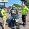 A imagem mostra um motociclista recebendo adesivo refletivo e ao lado o mascote da UGMT, Vaguinho.
