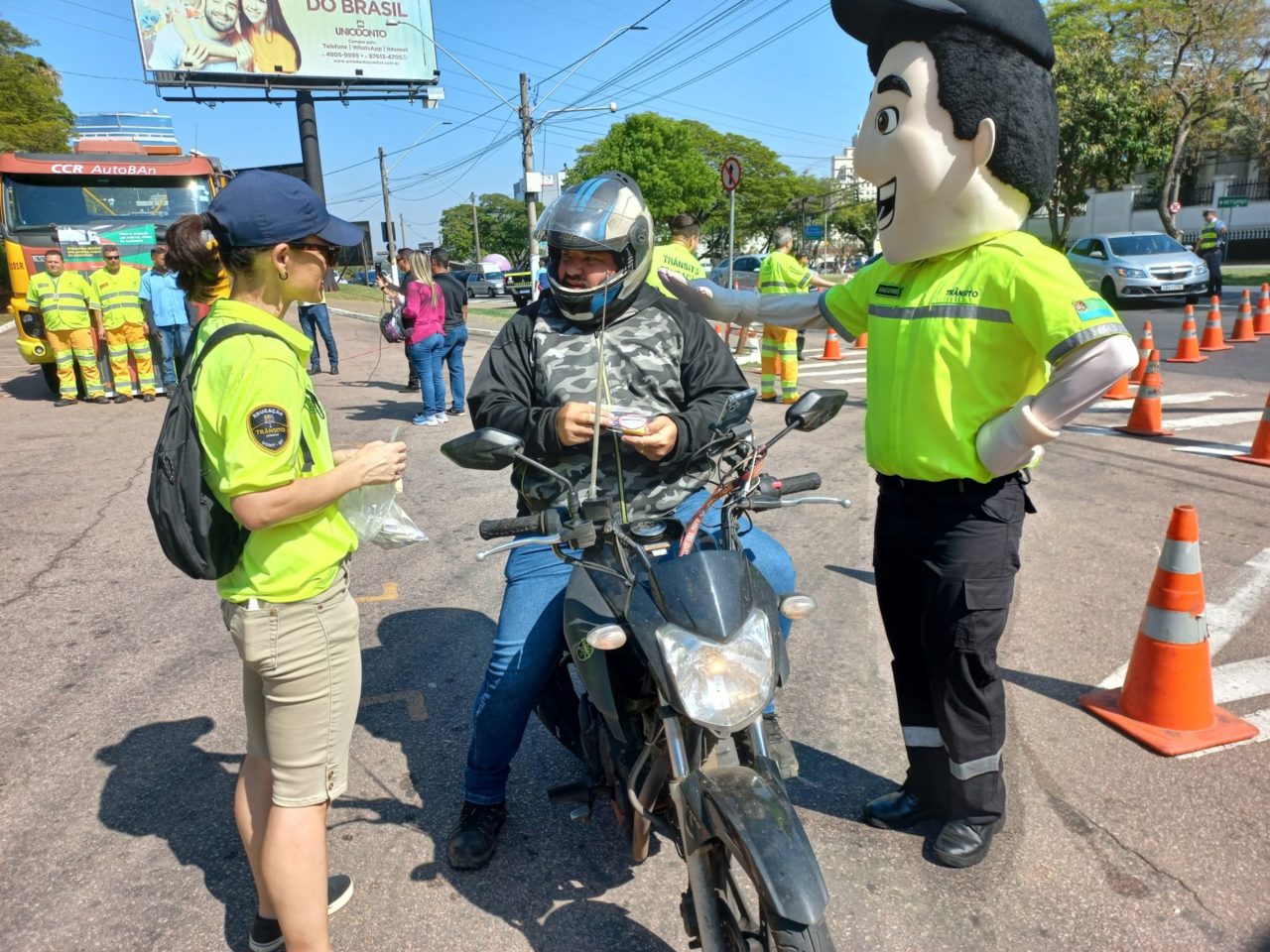 A imagem mostra um motociclista recebendo adesivo refletivo e ao lado o mascote da UGMT, Vaguinho.
