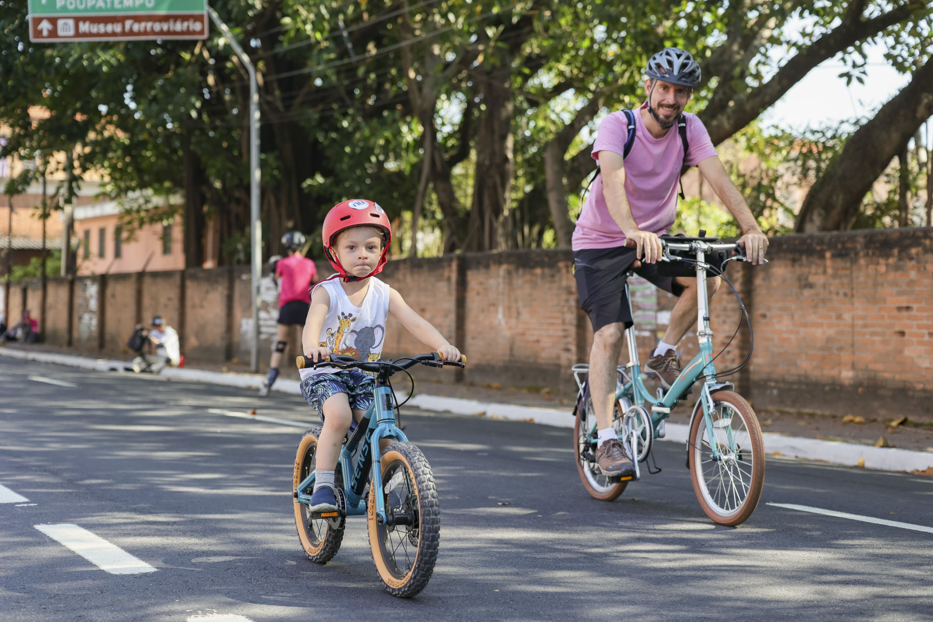 a imagem mostra um menino pequeno com capacete andando de bike ao lado do pai que também está de capacete e andando de bike. Evento Mobilidade Saudável e Lazer na Vila Arens e região Central neste domingo.