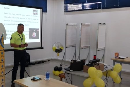 A imagem mostra um dos palestrantes da equipe de Educação para o Trânsito em uma empresa. Atrás dele há um telão com apresentação sobre o risco do uso de celulares quando se dirige.