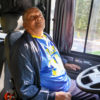 A imagem mostra um idoso sentado no lugar do motorista de ônibus, ele olha para o retrovisor para tentar enxergar uma pessoa no momento do embarque.