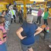 A imagem mostra um grupo de idosas fazendo atividade física, além de professoras e do mascote da UGMT, Vaguinho.