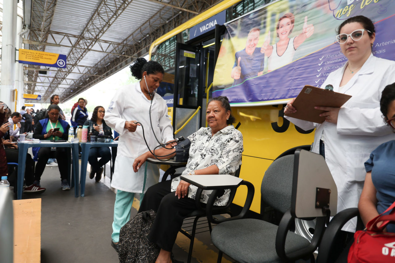 A imagem mostra uma mulher medindo a pressão arterial com uma enfermeira da Saúde da Prefeitura de Jundiaí. Elas estão na plataforma do Terminal Central.