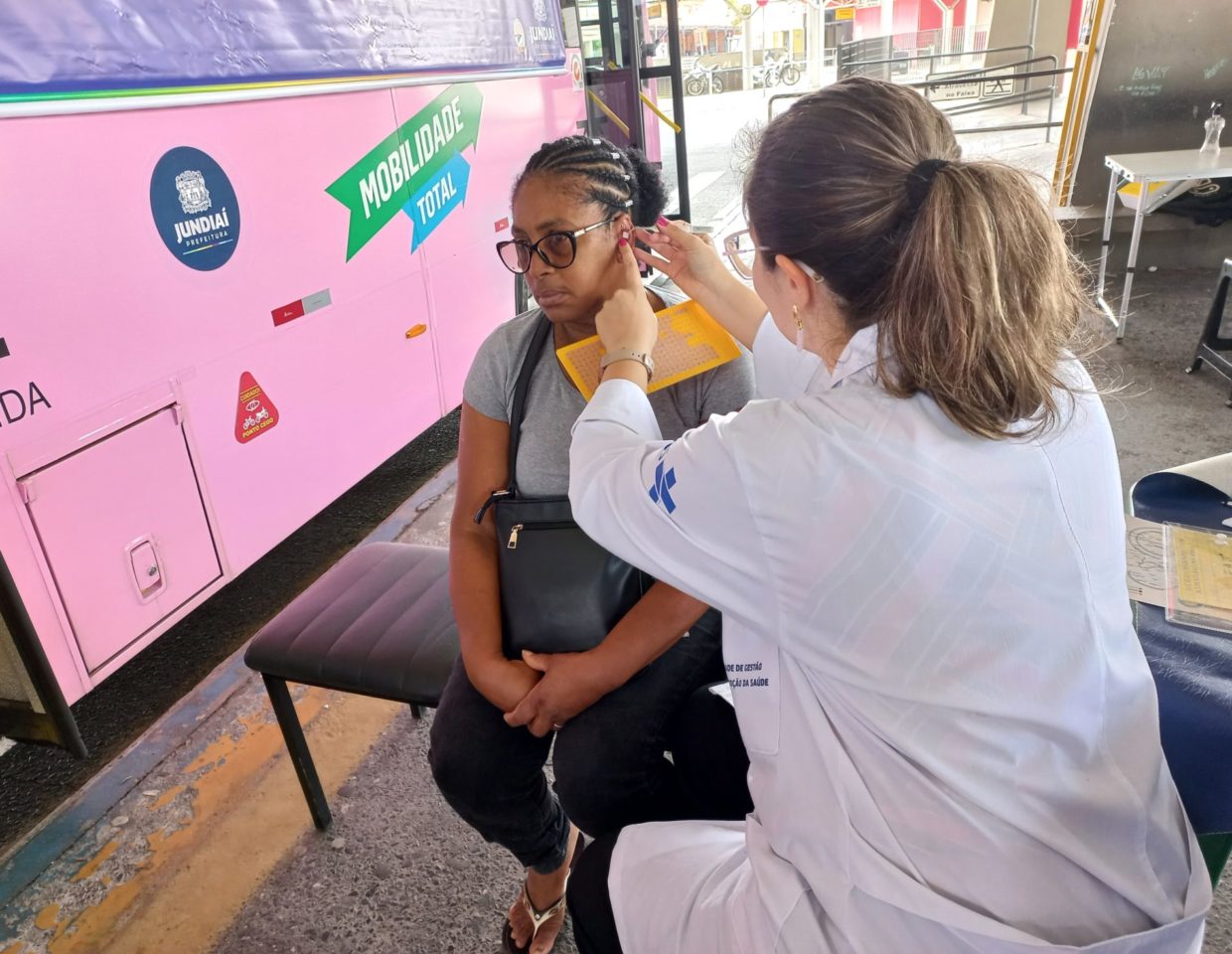 A imagem mostra uma enfermeira fazendo auriculoterapia em uma senhora na plataforma do Terminal Vila Arens.