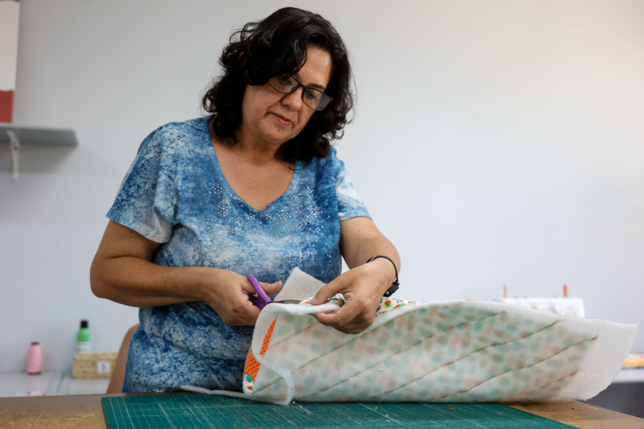 Senhora de cabelos pretos sentada, de óculos, cortando um pano com uma tesoura no curso de costura criativa.