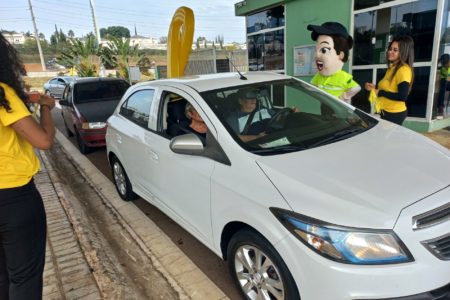 Na imagem, condutor e passageiros de um carro estão parados enquanto recebem orientações da mascote da Unidade de Gestão de Mobilidade e Transporte, na portaria do Parque da Cidade.