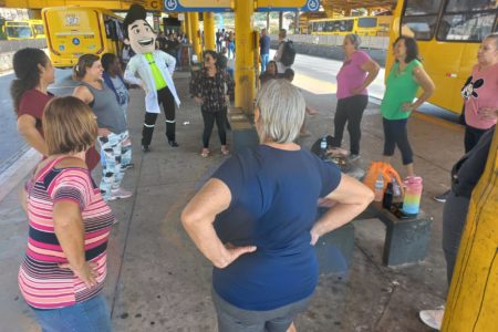 A imagem mostra um grupo de idosas fazendo atividade física, além de professoras e do mascote da UGMT, Vaguinho.